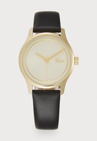 Reloj de tono dorado con esfera beige texturizada, correa de cuero negra y agujas doradas. Presenta una corona para ajustes y un diseño minimalista.