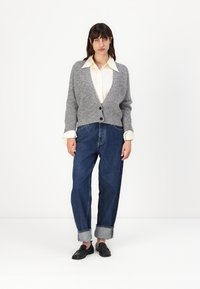 Cardigan grigio a maglia con scollatura a V profonda, chiusura con bottoni, indossato sopra una camicia bianca. Abbinato a jeans in denim blu a vita alta e larghi.