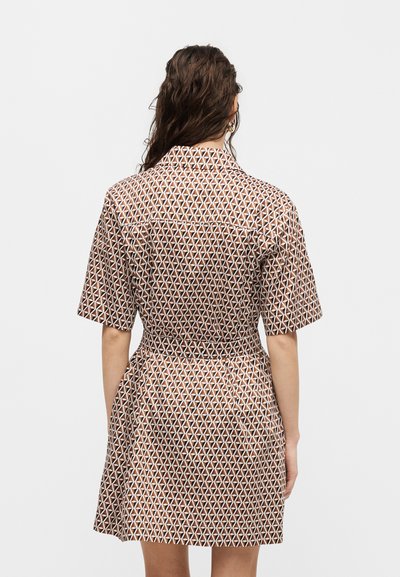 Γυναίκα με μακριά μαλλιά που φοράει ένα κοντομάνικο φόρεμα μέχρι το γόνατο με καφέ και άσπρο γεωμετρικό σχέδιο, που στέκεται με την πλάτη εμφανώς σε απλό φόντο.