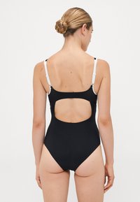 Donna con un chignon intrecciato che indossa un costume intero nero con spalline regolabili bianche e un grande taglio sulla schiena, in piedi su uno sfondo bianco.
