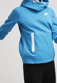 Blå Nike-huvtröja med vit dragkedjeficka, bärs av en person med armarna något utsträckta, i kombination med svarta byxor framför en enhetlig bakgrund.