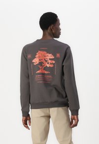 Grå sweatshirt med en röd bonsai-trädgrafik och text på baksidan, med ribbat halsband och ärmslut, tillverkad av ett mjukt material.