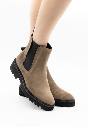 Knöchelhohe taupefarbene Wildlederstiefel mit schwarzen elastischen Seitenpaneelen und dicken Profilsohlen, getragen an bloßen Beinen.