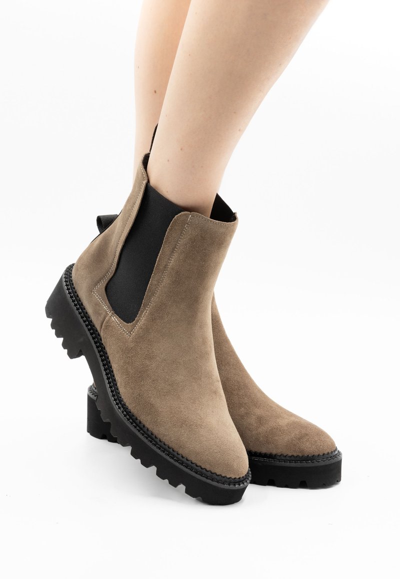 Bottes en daim taupe longueur cheville avec panneaux latéraux élastiques noirs et semelles épaisses à crampons portées sur jambes nues.