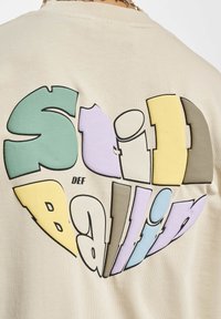 Beige bomulds T-shirt med et stort hjerteformet grafisk motiv. Tekst i farverige fede skrifttyper: "Sliid Ballin" med "def" i mindre sort skrifttype.