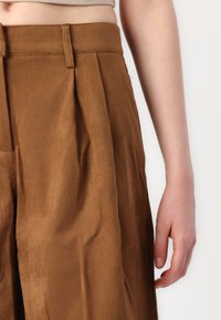 Pantalons en daim marron avec un design plissé et une taille structurée. Texture lisse, montrée avec une main reposant sur le tissu.