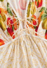 Close-up van stof met oranje en groene bloemenprint en gele decoratieve print, met twee gebonden witte koordjes met zilveren uiteinden.