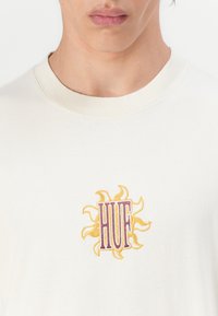 Kremfarget bomull t-skjorte med et brodert sol-design i gult og lilla, med bokstavene "HUF" i midten.