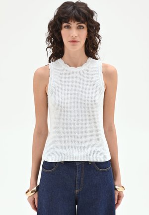 Donna con capelli ricci scuri che indossa un top bianco senza maniche in maglia e jeans blu, in piedi davanti a uno sfondo chiaro e uniforme.