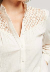Chemise beige clair avec yoke en tulle brodé de fleurs, devant à boutons et corsage plissé. Manches longues avec détails de manchette. Texture douce.