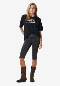 Juodas per didelis T-shirt su "oasis" logotipu, derinamas su tamsiai pilkais džinsiniais šortais ir rudos spalvos kelių ilgio botais. Paprastas, kasdienis stilius.