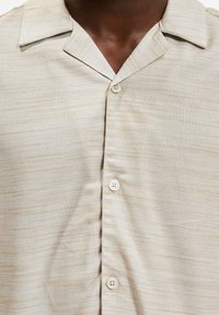 Lysærmet beige skjorte med teksturert stoff, med en klippet krage og to hvite knapper foran. Svake diagonale striper til stede.