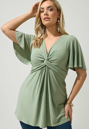 Femme blonde portant un chemisier vert sauge à nœud torsadé en V avec manches papillon, boucles d'oreilles en forme de feuille dorée et bracelet en or, posant devant un fond clair.