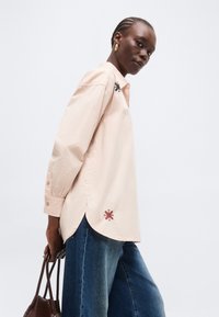 Γυναίκα με μπεζ πουκάμισο μακρυμάνικο με κεντητά μοτίβα, μπλε τζιν, κρατώντας καφέ υφασμάτινη τσάντα, κοιτάζει προς την κάμερα.