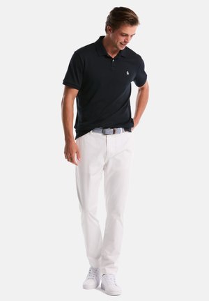 Polo bleu marine avec un col classique, des manches courtes et un logo, associé à un pantalon blanc et des baskets blanches, créant un look décontracté.