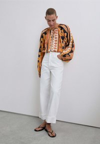 Blusa de mangas largas a rayas en naranja y negro, pantalones blancos de tiro alto y pierna ancha, y chanclas negras con detalles de tachuelas.