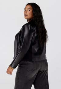 Zwarte leren jas met een getailleerd ontwerp, ritssluiting aan de voorkant enTextuurrugaccenten; gecombineerd met donkergrijze jeans.