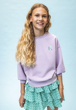 Lila sweatshirt met een ribbelboord en turquoise geborduurde "K," gecombineerd met een groene rok met ruches en een patroon. Golvend haar.