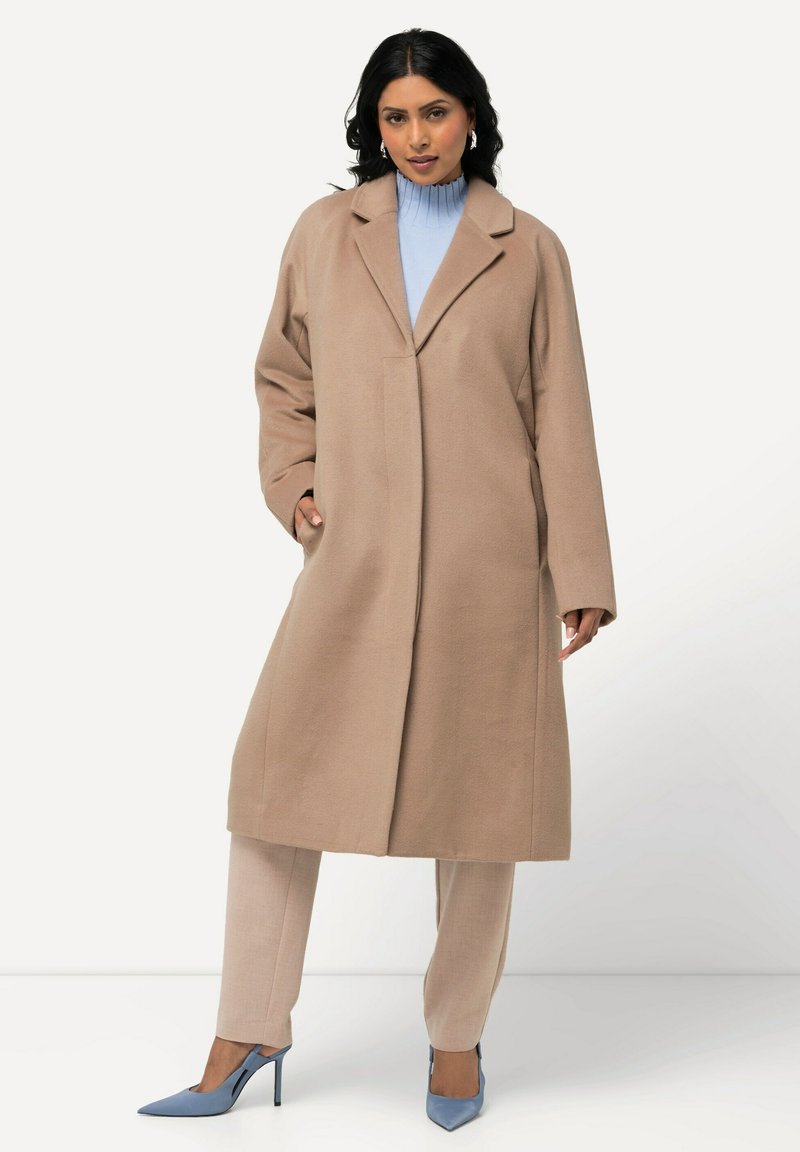 Cappotto lungo beige con rever, tasche laterali e vestibilità ampia, abbinato a un top azzurro chiaro, pantaloni beige e tacchi blu.