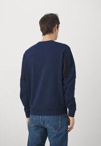 Felpa blu navy con colletto tondo, pannelli in rete nera testurizzata sulle spalle e polsini a costine. Indossata con pantaloni di jeans blu.
