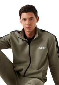 Chándal verde oliva con cremallera y una franja negra en las mangas, con el logo "GUESS" en blanco. Textura suave, ajuste atlético y cuello alto.