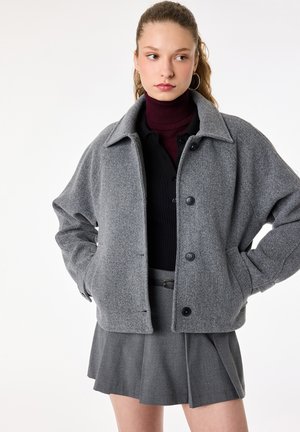 Joven mujer con las manos en los bolsillos, vistiendo una chaqueta de lana gris abotonada, top negro, jersey de cuello alto burdeos y falda gris plisada.