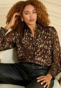 Blusa marrone con stampa leopardata metallica, design con bottoni e polsini plissettati; abbinata a pantaloni neri aderenti. Texture liscia.