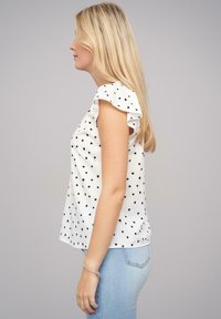 Weiße Bluse mit schwarzen Herzmustern, Rüschensäumen und lockerer Passform. Kombiniert mit hellblauen Jeans. Glatte Stofftextur.