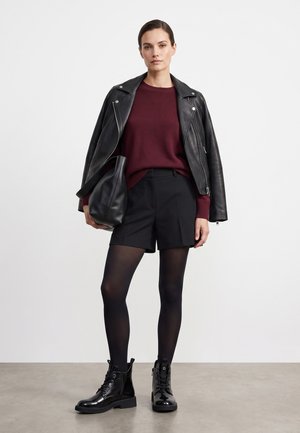 Kvinde iført en sort læderjakke over en bordeauxrød sweater, sorte shorts, gennemsigtige sorte strømpebukser og sorte snørestøvler til ankler.