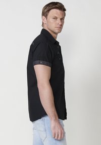 Camisa negra de manga corta con cuello abotonado, con puños en contraste y sutiles detalles bordados. La tela parece ligera y estructurada.