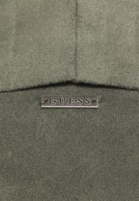 Tessuto verde oliva con una texture morbida, caratterizzato da un'etichetta in metallo con il logo "GUESS". Il design include un colletto e dettagli di cucitura.