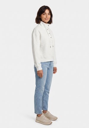 Donna in piedi che indossa un pullover trapuntato bianco con bottoni a pressione, jeans azzurri e scarpe da ginnastica beige su sfondo bianco.