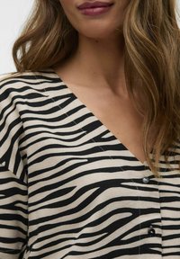 Haut rayé noir et beige avec un décolleté en V, trois boutons et un tissu léger et texturé.