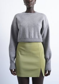 Personne portant un pull gris à manches longues et une mini-jupe vert clair avec une fente à l'avant, debout devant un fond blanc uni.