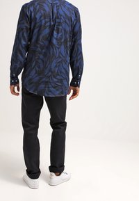 Mężczyzna ubrany w niebieską koszulę w wzory z długimi rękawami, czarne spodnie i białe trampki, stojący tyłem na jednolitym jasnym tle.