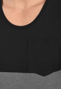 Sort tanktop med en grå vandret stribe. Har en lomme på venstre bryst og en rund halsudskæring. Fremstillet af blødt bomuldsstof.