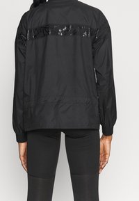 Schwarze Windjacke mit einem glänzenden schwarzen Paneel am oberen Rücken; verfügt über elastische Bündchen und einen Kordelzugsaum.