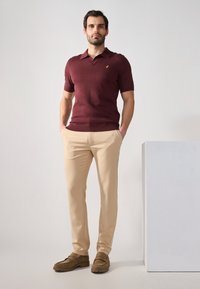 Burgundin lyhythihainen polo-paita kauluksella, beige housut ja ruskeat mokkasiinit; paidassa on pieni logo rinnassa.
