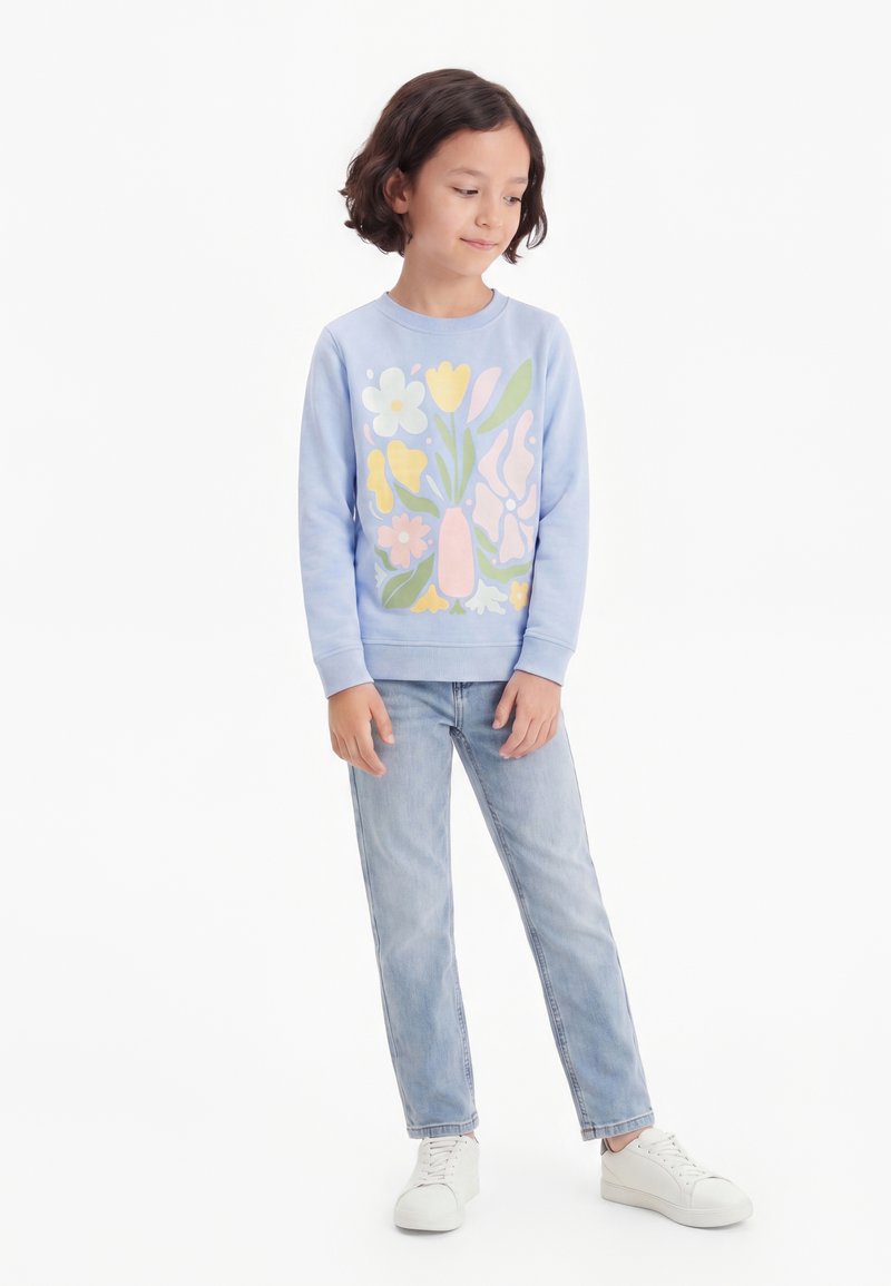 Enfant portant un sweat-shirt bleu clair avec un motif floral coloré, un jean délavé clair et des baskets blanches, debout devant un fond blanc.