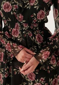 Abito floreale con tessuto nero trasparente e motivi di rose rosa, vita arricciata e maniche lunghe trasparenti. Mani posizionate vicino all'orlo.