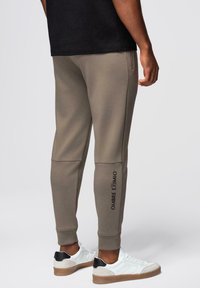 Mężczyzna w taupe joggerach z czarnym napisem "OMBRE EXIMIO" na dolnej nodze i białych sneakersach z czarnymi akcentami na pięcie, stojący na tle szarym.