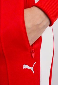 Crvena sportska jakna s patentnim zatvaračem i džepom, s bijelim bočnim panelima i teksturiranim materijalom. Uključuje mali bijeli Puma logotip na džepu.