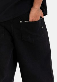 Pantalones de mezclilla negros con un ajuste relajado. Presentan bolsillos laterales con herrajes plateados y una etiqueta de marca visible en el bolsillo. Textura suave.
