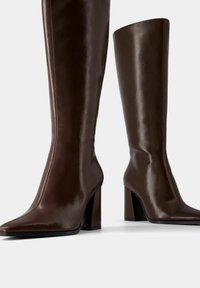 Para wysokich, ciemnobrązowych skórzanych kozaków z ostro zakończonymi noskami i masywnymi wysokimi obcasami na białym tle.