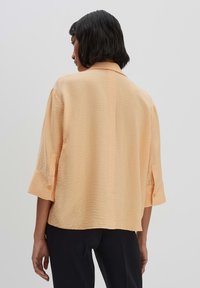 Blusa leggera di colore pesca con finitura testa di moro, dotata di colletto standard e maniche arrotolate. I bottoni decorano i polsini e il retro.
