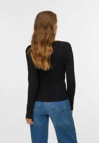 Donna con lunghi capelli ondulati castano chiaro, che indossa una maglietta nera a maniche lunghe e jeans blu, in piedi di spalle contro uno sfondo semplice.