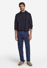 Marinblå tröja med huva, dragen över en mörk polotröja, kombinerad med blå jeans och beige sneakers, vilket skapar en avslappnad stil.