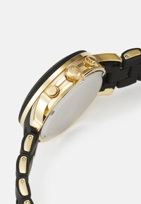 Reloj en tono dorado con una correa de silicona negra. La caja tiene dos coronas y botones dorados, con una esfera redonda y suave.