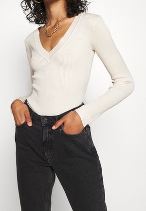 Persoon draagt een geribde crèmekleurige V-hals trui en zwarte high-waist jeans met handen in de zakken tegen een witte achtergrond.