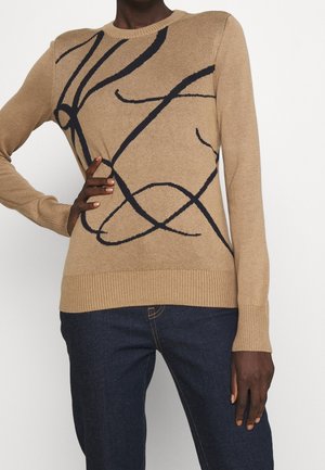 Pull beige à manches longues avec des lignes abstraites noires, associé à un jean bleu foncé, porté par une personne avec la main sur la taille et une posture détendue.